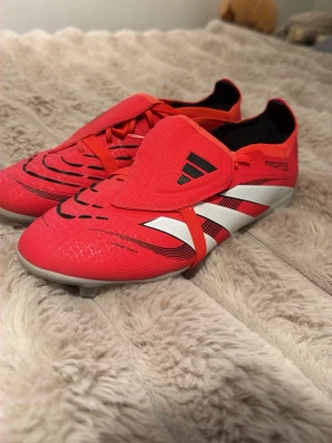 Adidas Predator röda fotbollsskor barn elite - Säljer ett par Adidas Predator fotbollsskor i rött med svarta och vita detaljer. Skorna har klassiska tre ränder på sidan, snörning och täckt plös. Yttersulan är vit med svart text och dobbar för bra grepp på planen. Materialet är syntet för lätt vikt och bra bollkontroll.