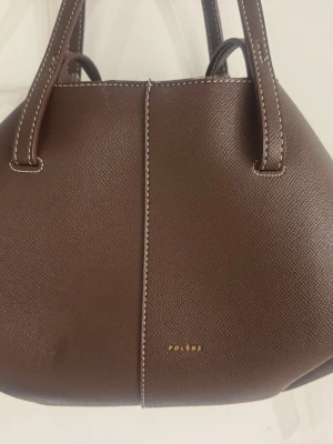 Brun handväska från Polène - Snygg brun handväska från Polène i strukturerat läder. Väskan har en unik, geometrisk form med dubbla handtag och kontrastsömmar. Perfekt för dig som vill ha en stilren och modern accessoar till din outfit.