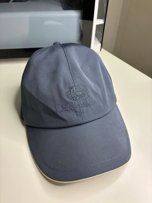 Blå keps från Loro Piana - Snygg blå keps från Loro Piana med broderad logga framtill. Klassisk böjd skärm och justerbar passform bak. Tillverkad i ett mjukt tyg som ger en clean och stilren look. Perfekt accessoar för att lyfta din outfit.