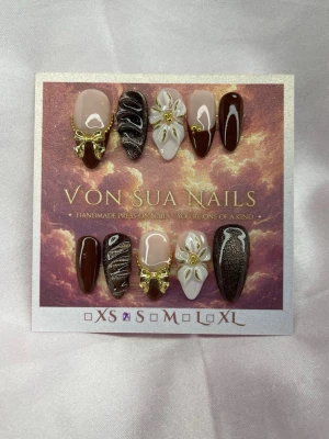 ChocoCouture (S);Medium Almond - Snygga handgjorda press-on naglar i en medium lång mandelform med mix av bruna,cateye, vita french och nude bas. Designen har glittercateye, 3D-blommor och guldfärgade rosetter för en lyxig känsla. Alla naglarna är handgjorda och ett prep-kit skickas med. Perfekt för dig som vill ha något unikt och trendigt.S: 15 mm, 11,5 mm, 12 mm, 11 mm, 9 mm 