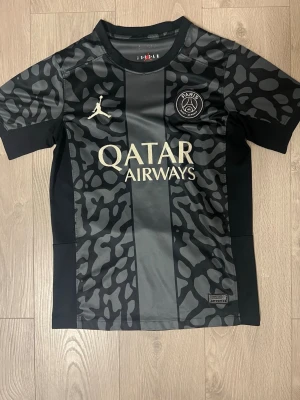 Svart PSG x Jordan matchtröja - Snygg svart PSG matchtröja i samarbete med Jordan. Tröjan har ett unikt camomönster, PSG-logga på bröstet och Jordan-logga i vitt. Qatar Airways är tryckt i vitt framtill. Tillverkad i ett lätt och ventilerande material, perfekt för fotboll eller träning.