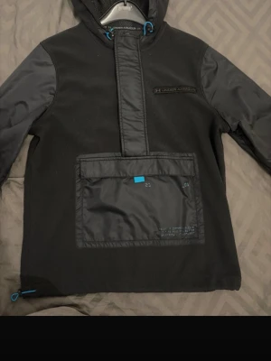 Svart under armour vindjacka  - Snygg svart vindjacka från Under Armour med huva och stora fickor framtill. Jackan har detaljer i nylon på ärmar och ficka, samt blå dragsko vid huvan och nederkanten. Perfekt för blåsiga dagar och streetwear-stil.