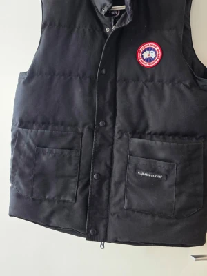 Svart dunväst från Canada Goose - Svart dunväst från Canada Goose med klassisk logotyp på bröstet och två stora fickor framtill. Västen har tryckknappar och hög krage, perfekt för att hålla värmen. Materialet är slitstarkt och fyllningen är dun, vilket gör den riktigt varm och bekväm.
