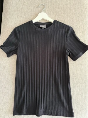Svart ribbad t-shirt - Snygg svart t-shirt med ribbad struktur och rund hals. T-shirten har korta ärmar och en stilren look som passar till det mesta. Tillverkad i ett mjukt material som känns skönt mot huden. Perfekt för dig som gillar enkel men trendig stil.