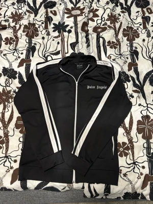 Svart hoodie från Palm Angels M - Svart hoodie från Palm Angels med vita ränder längs ärmarna och dragkedja framtill. Klassisk streetwear-look med logga broderad på bröstet. Tillverkad i ett mjukt och stretchigt material som passar perfekt till en avslappnad stil.