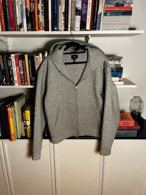 Grå zip-hoodie med huva - Snygg grå hoodie med dragkedja och huva. Klassisk modell med två fickor framtill och ribbade muddar vid ärmslut och nederkant. Perfekt för en avslappnad och stilren look.