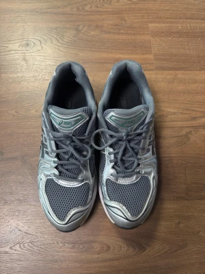 Gråa Asics sneakers med meshdetaljer - Snygga grå sneakers från Asics med silvriga detaljer och meshpaneler för extra ventilation. Skorna har klassisk snörning och en sportig look som passar perfekt till streetwear eller träning. Tidlös design med bekväm vadderad insida.