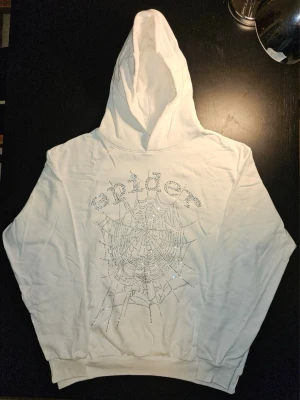 Vit Sp5der Rhinestone Web Hoodie - Vit hoodie med stort spindelnätsmotiv och glittrande rhinestones framtill. Snygg oversized/streetwear-look med känguruficka och huva. Helt oanvänd och i nyskick. Förvarad i skyddspåse. Se bilder för detaljer och skick.