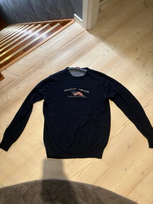 Mörkblå Gant tröja med tryck - Mörkblå långärmad tröja från Gant med rund halsringning och tryck framtill med texten 'GANT 1949' samt en amerikansk flagga. Tröjan har en klassisk passform och ribbade muddar vid ärmslut och nederkant.