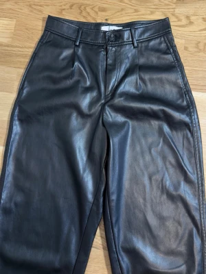 Mango svarta byxor i skinnimitation – storlek 36/S - 🖤 Stilrena svarta byxor i mjuk skinnimitation ✨ Hög midja med veck framtill 👖 Rak, lätt avsmalnande passform 📌 Sidfickor och hällor för skärp 🏷️ Storlek 36/S  Material: 100 % polyuretan. Foder: 100 % polyester.  Mått finns på bilderna. Kontrollera gärna måtten före köp. Jag erbjuder mängdrabatt vid köp av flera varor. Se gärna mina andra annonser.  