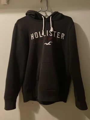 Svart hoodie från Hollister - Svart hoodie från Hollister med vit logga och röd detalj på bröstet. Klassisk känguruficka framtill, vita dragsnören i huvan och mjukt material. Perfekt för en chill och avslappnad stil.