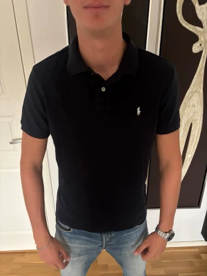 Svart pikétröja från Polo Ralph Lauren - Klassisk mörkblå pikétröja från Polo Ralph Lauren med vit broderad logga på bröstet. Tröjan har krage, knappar vid halsen och korta ärmar. Tillverkad i mjuk bomull som känns skön mot huden. Perfekt till jeans eller shorts. Storlek L och mycket bra skick.