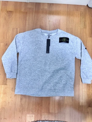 Ljusgrå sweatshirt från Stone Island - Ljusgrå sweatshirt från Stone Island med klassisk rund hals och ribbade muddar. Tröjan har den ikoniska Stone Island-loggan som kan fästas på ärmen. Perfekt för en avslappnad och stilren look.