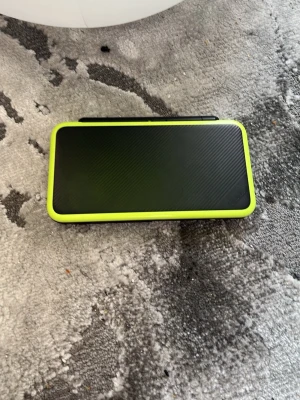 (Nintendo)Svart och limegrön bärbar spelkonsol - 2DS XL Nintendo Säljer en bärbar spelkonsol i svart med limegröna detaljer. Konsolen har vikbar design med två skärmar, styrkors och knappar i limegrönt. Perfekt för gaming på språng och enkel att ta med sig tack vare det smidiga formatet.