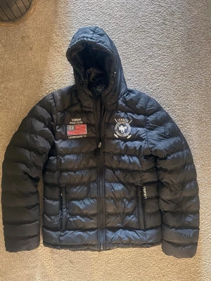 Svart pufferjacka från Vinson Polo Club - Snygg mörkblå pufferjacka från Vinson Polo Club med huva och dragkedja. Jackan har broderade detaljer på bröstet, inklusive klubbens logga och amerikansk flagga. Tillverkad i glansigt, quiltat material som håller värmen under kalla dagar. I fint skick och varm och skön 