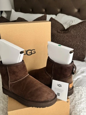 Bruna UGG moon boots i mocka - Säljer ett par klassiska bruna UGG moon boots i mjuk mocka med fårskinnsfoder. De har en rund tå, låg skaft och grov sula för extra komfort. Perfekta för kalla dagar och riktigt sköna att ha på sig. De kan ha tecken på användning då materialet är lätt slitsamt men fortfarande väldigt bra kvalitet. De säljs i kartong som på bild