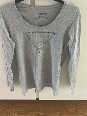 Grå långärmad topp från Guess - Snygg grå långärmad topp från Guess med rund halsringning. Framtill finns det klassiska Guess-triangelloggan dekorerad med små glittriga stenar. Perfekt till jeans eller kjol för en clean och stilren look.