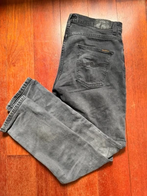 Grå jeans från Nudie Jeans Co - Säljer ett par grå jeans från Nudie Jeans Co med klassisk femficksmodell och raka ben. Jeansen har en diskret logga på bakfickan och är tillverkade i mjuk bomull. Perfekta för en avslappnad och stilren look.