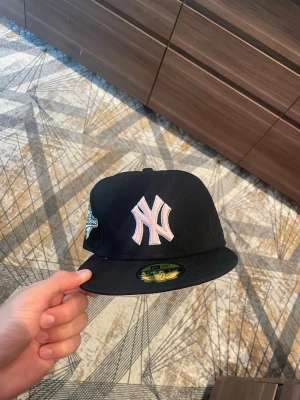 Svart New Era Yankees keps - Snygg svart 59FIFTY keps från New Era med New York Yankees-logga i vitt och rosa framtill. Klassisk flat brim och broderad detalj på sidan. Perfekt för dig som vill ha en stilren och sportig look.