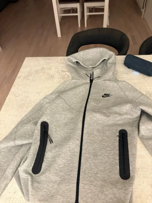 Grå Nike Tech hoodie med svarta detaljer - Snygg grå hoodie från Nike med svarta dragkedjor på fickorna och svart Nike-logga på bröstet. Hoodien har hel dragkedja framtill och en stor huva. Perfekt för en sportig och avslappnad stil.