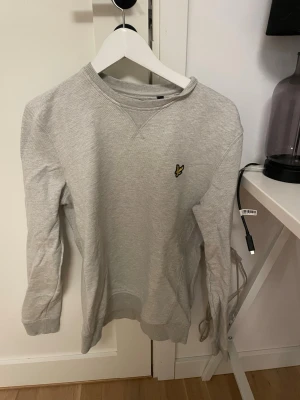 Grå sweatshirt från Lyle & Scott - Klassisk grå sweatshirt från Lyle & Scott med rund halsringning och ribbade muddar. Tröjan har den ikoniska gula örnloggan broderad på bröstet och är tillverkad i mjukt bomullsmaterial. Perfekt för en avslappnad och stilren look.
