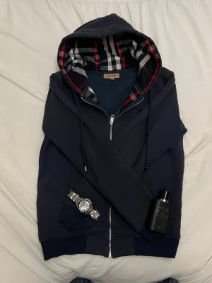 Marinblå hoodie från Burberry - Snygg marinblå hoodie från Burberry med dragkedja och klassiskt rutigt foder i huvan. Hoodien har dragsko vid huvan och ribbade muddar. Perfekt för dig som vill ha en stilren och exklusiv look.