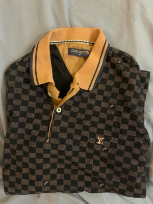 Rutig pikétröja från Louis Vuitton - Snygg pikétröja från Louis Vuitton med klassiskt rutigt mönster i svart och beige. Tröjan har en gul krage med blå ränder och knappslå framtill. LV-logga broderad på bröstet. Perfekt för dig som vill sticka ut med lyxig stil.