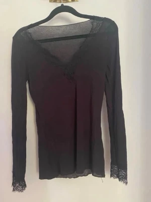 Svart långärmad topp från Intimissimi - Svart långärmad topp från Intimissimi i ultralätt material med kashmir. V-ringad med spetsdetaljer vid halsringning och ärmslut. Skriv om bild på 