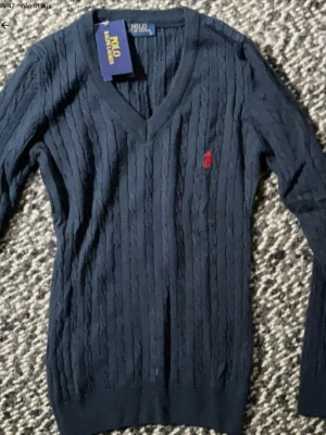 Mörkblå kabelstickad tröja Polo Ralph Lauren - Kabelstickad mörkblå tröja från Polo Ralph Lauren med V-ringning och röd broderad logga på bröstet. Tröjan har långa ärmar och ribbade muddar. Perfekt för dig som gillar klassisk och stilren design.