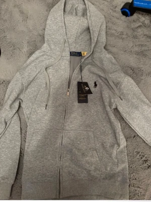 Grå hoodie från Polo Ralph Lauren - Snygg grå hoodie med dragkedja från Polo Ralph Lauren. Klassisk design med huva och justerbara snören, samt fickor framtill. Liten broderad logga på bröstet. Tillverkad i mjukt bomullsmaterial som känns skönt mot huden.