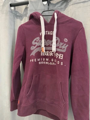 Vinröd hoodie från Superdry - Säljer en vinröd hoodie från Superdry med vit logga och text framtill. Hoodien har stor känguruficka, vita grova dragsnören och ribbade muddar. Perfekt för dig som gillar streetwear och vill ha något bekvämt och snyggt.