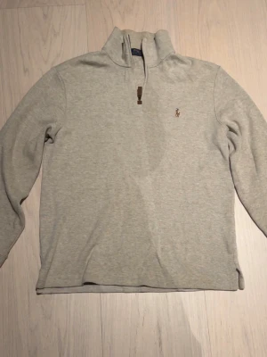 Ljusgrå tröja Polo - Ljusgrå långärmad tröja från Polo Ralph Lauren med halv dragkedja och liten broderad logga på bröstet. Tröjan har hög krage och är tillverkad i mjukt material, perfekt för lager-på-lager-stil.