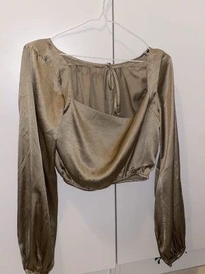 Beige blus - Snygg champagnefärgad blus i glansigt satinmaterial med draperad front och resår i midjan. Blusen har långa ärmar med lätt puff och knytning i ryggen. Perfekt för en trendig och elegant look.