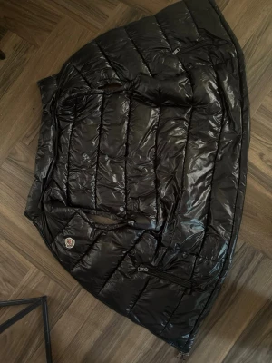 Moncler black polyamid - Snygg svart dunväst med glansig finish och horisontella sömmar. Västen har två framfickor med dragkedja och en rund patch på bröstet. Perfekt för lager-på-lager och ger en modern look till din outfit.