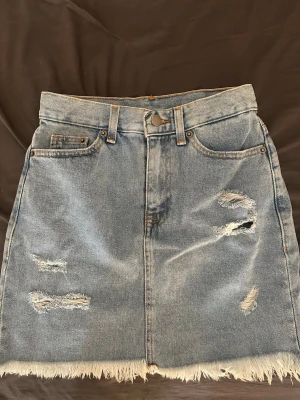 Blå jeanskjol med slitningar - Säljer en kort blå jeanskjol med slitna detaljer och fransig nederkant. Kjolen har klassiska fem fickor, bälteshällor och knappstängning framtill. Perfekt för dig som gillar en avslappnad och trendig stil.