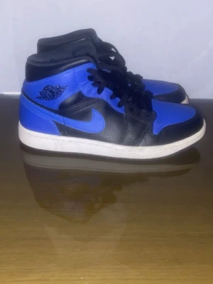 Air Jordan 1 Mid Svart/Blå - Snygga Air Jordan 1 Mid sneakers i svart och blått läder med klassisk vit sula. Skorna har svarta skosnören, ikonisk Swoosh och Jordan Wings-logga på sidan. Perfekta för dig som gillar streetwear och vill sticka ut med en färgstark look.