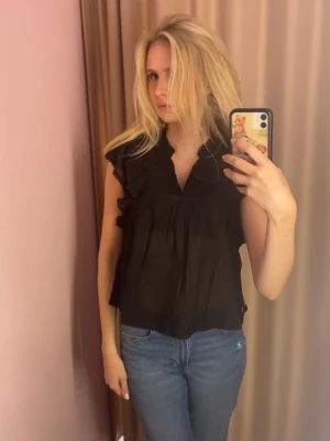 Svart volangblus med broderi - Neo noir liknande. Säljer en svart blus med volangdetaljer på axlarna och v-ringning. Blusen har broderade små prickar i tyget och smockdetalj över bröstet. Luftig och lätt modell som ger en avslappnad men ändå chic look.