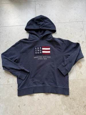 Mörkblå hoodie från Lexington - Mörkblå hoodie från Lexington med broderad amerikansk flagga och texten 'American Heritage Since 1993' på bröstet. Klassisk huva och ribbade muddar. Perfekt för en avslappnad och stilren look.