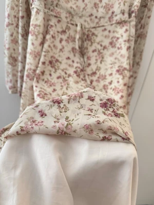 Blommig maxiklänning i chiffong med knytband - Söt och romantisk maxiklänning i ett ljusbeige/creme färgat chiffongtyg med ett subtilt rosa och lila blommönster. Klänningen har sweetheart-halsringning, smickrande avskärning under bysten, tunt knytband i midjan och långa ärmar med resår i ärmsluten. Helt fodrad för bekväm passform. Perfekt för sommarfester, bröllop eller en finare vardagsstil.