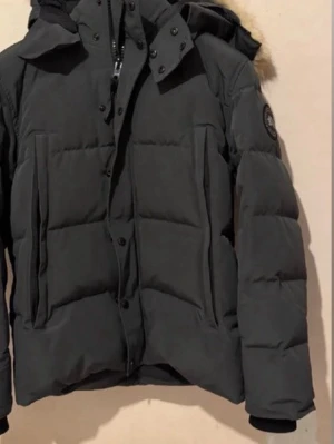 Svart dunjacka med pälskantad huva - Snygg svart dunjacka med stor huva och pälskant. Jackan har quiltad design, två stora fickor framtill och ribbade muddar vid ärmsluten. Märkespatch på ena ärmen ger extra stilpoäng. Perfekt för kalla vinterdagar.