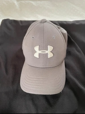 Grå keps från Under Armour - Snygg grå keps från Under Armour med vit broderad logga framtill. Kepsen har böjd skärm och är tillverkad i ett ventilerande meshmaterial som ger en sportig look.
