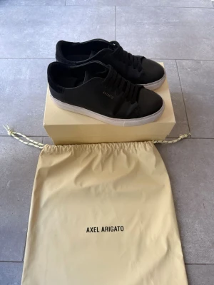 Svarta Axel Arigato sneakers - Snygga svarta sneakers från Axel Arigato med minimalistisk design och gulddetalj på sidan. Skorna har vit sula och är tillverkade i läder med textildetaljer vid hälen. I bra skick och väldigt sparsamt användas. Dock en defekt,  se bild. Nypris 2290kr 