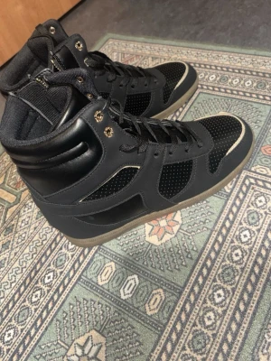 Svarta high top sneakers med meshdetaljer - Säljer ett par svarta high top sneakers med snygga meshdetaljer och svarta skosnören. Skorna har en gummisula i beige och metallfärgade öljetter. Materialet är en mix av syntet och textil, vilket ger en sportig och modern look.