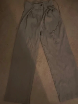 Beiga vida linnebyxor med knytband - Snygga beiga linnebyxor med hög midja och vida ben. Byxorna har ett knytband i midjan och stängs med knapp och dragkedja. Perfekta för en avslappnad och trendig look. Tillverkade i ett luftigt linnematerial.