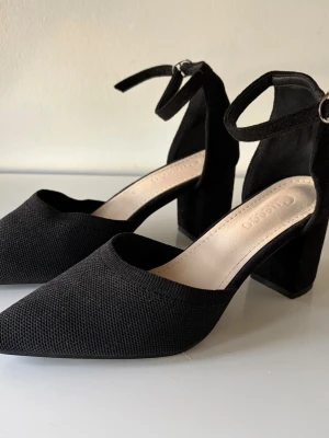 Svarta pumps med ankelrem och blockklack - Stilrena svarta pumps med spetsig tå, bred 7cm blockklack och justerbar ankelrem. Ovandelen är i textil med diskret struktur och klacken är klädd i mockaliknande material. Perfekta för dig som gillar en klassisk men trendig look. Oerhört sparsamt använda! Ärliga och snabba affärer uppskattas:)