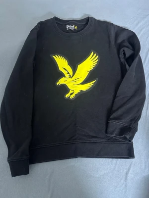 Svart sweatshirt från Lyle & Scott - Cool svart sweatshirt från Lyle & Scott med stor gul örn på bröstet. Tröjan har rund halsringning och ribbade muddar vid ärmslut och nederkant. Perfekt för en avslappnad och trendig look.