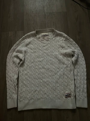 Vit kabelstickad tröja - Kabelstickad vit tröja med rund halsringning och ribbade muddar vid ärmslut och nederkant. Tröjan har en patch nertill på framsidan och ett klassiskt, tidlöst mönster. Perfekt för lager på lager under kyliga dagar.