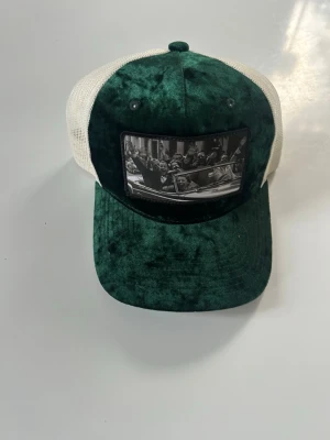 Grön och vit keps med tryck - Snygg keps i mörkgrön sammet med vit mesh baktill. Framtill finns ett svartvitt tryck med motiv av personer i en bil. Justerbar storlek med plastspänne. Perfekt accessoar för en cool streetstil.