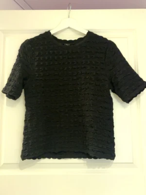 Svart strukturerad topp från Lindex - Cool svart topp från Lindex med unik strukturerad yta och korta ärmar. Toppen har en rund halsringning och är tillverkad i ett mjukt material som känns bekvämt mot huden. Perfekt för dig som vill ha något annorlunda i garderoben.