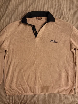 Beige stickad tröja med mörk krage - Säljer en beige stickad tröja med mörkblå krage och knappslå. Tröjan har lång ärm, ribbade muddar och en broderad logga på bröstet. Perfekt för dig som gillar klassisk och avslappnad stil.